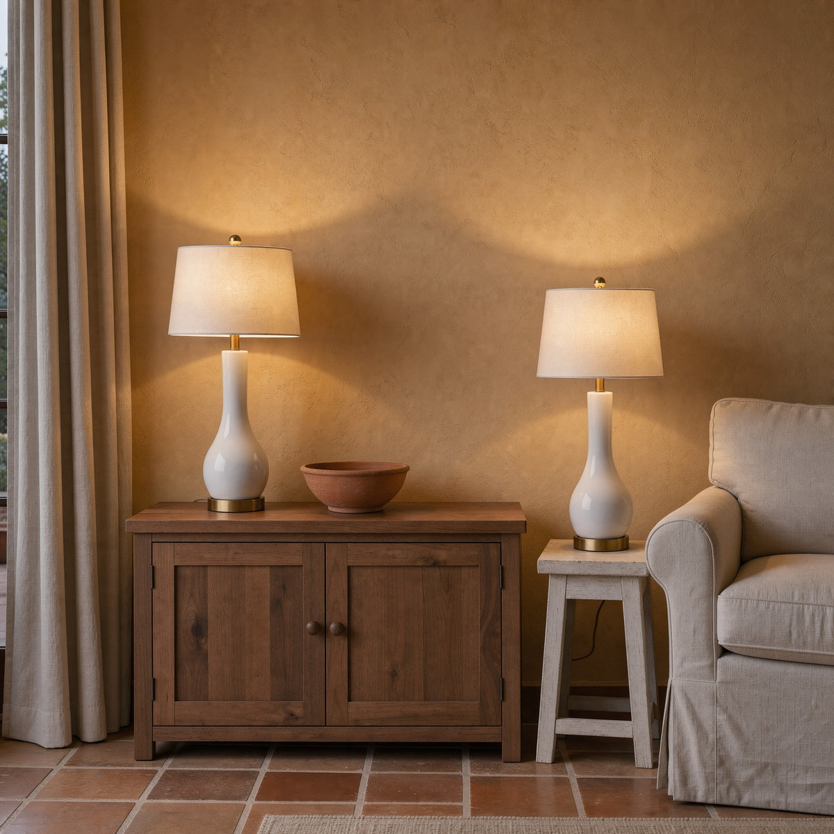 Pair of white ceramic table lamps with brass feet and white fabric shades, lit on a wood cabinet and a stool