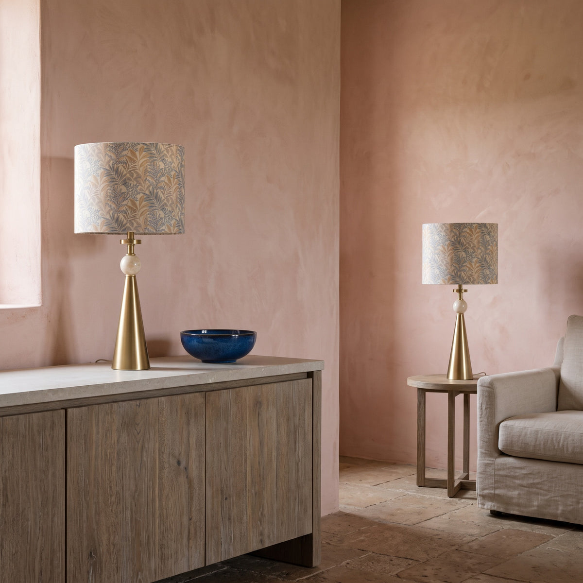 Pair of gold cone table lamps with travertine accents and leaf-print drum shades on an oak sideboard and side table