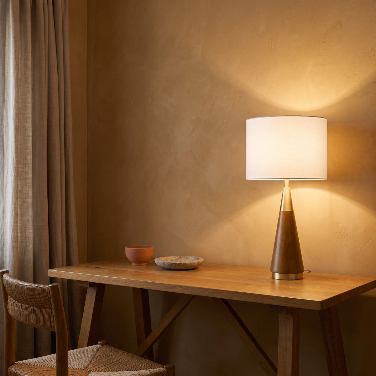 Wood and brass cone table lamp with white drum shade lit on an oak console beside a linen curtain