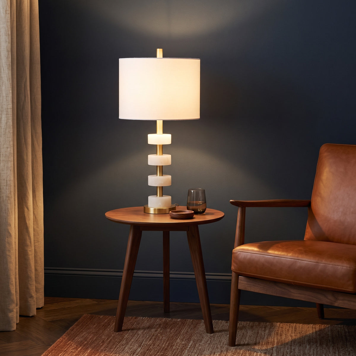 White marble and brass stacked disc table lamp, lit, on a round walnut side table beside a leather lounge chair