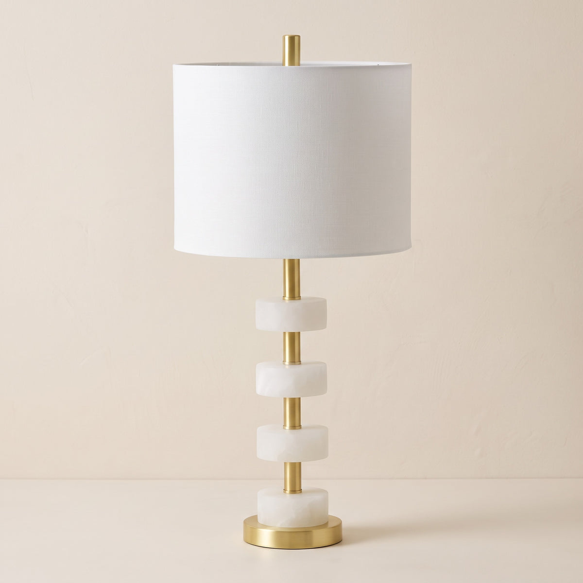 White marble stacked disc table lamp with brass stem and white drum shade on an ivory backdrop