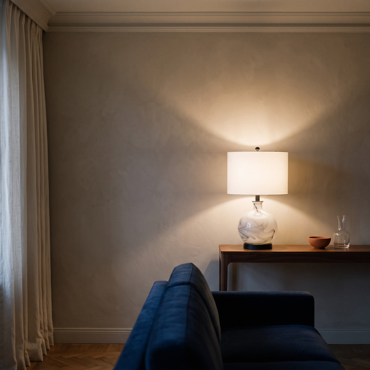 White and gray swirled glass table lamp, lit, with white drum shade on a walnut console beside a navy sofa