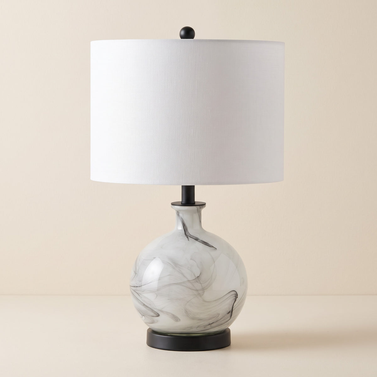 White and gray swirled glass table lamp with white linen drum shade and black base on an ivory backdrop