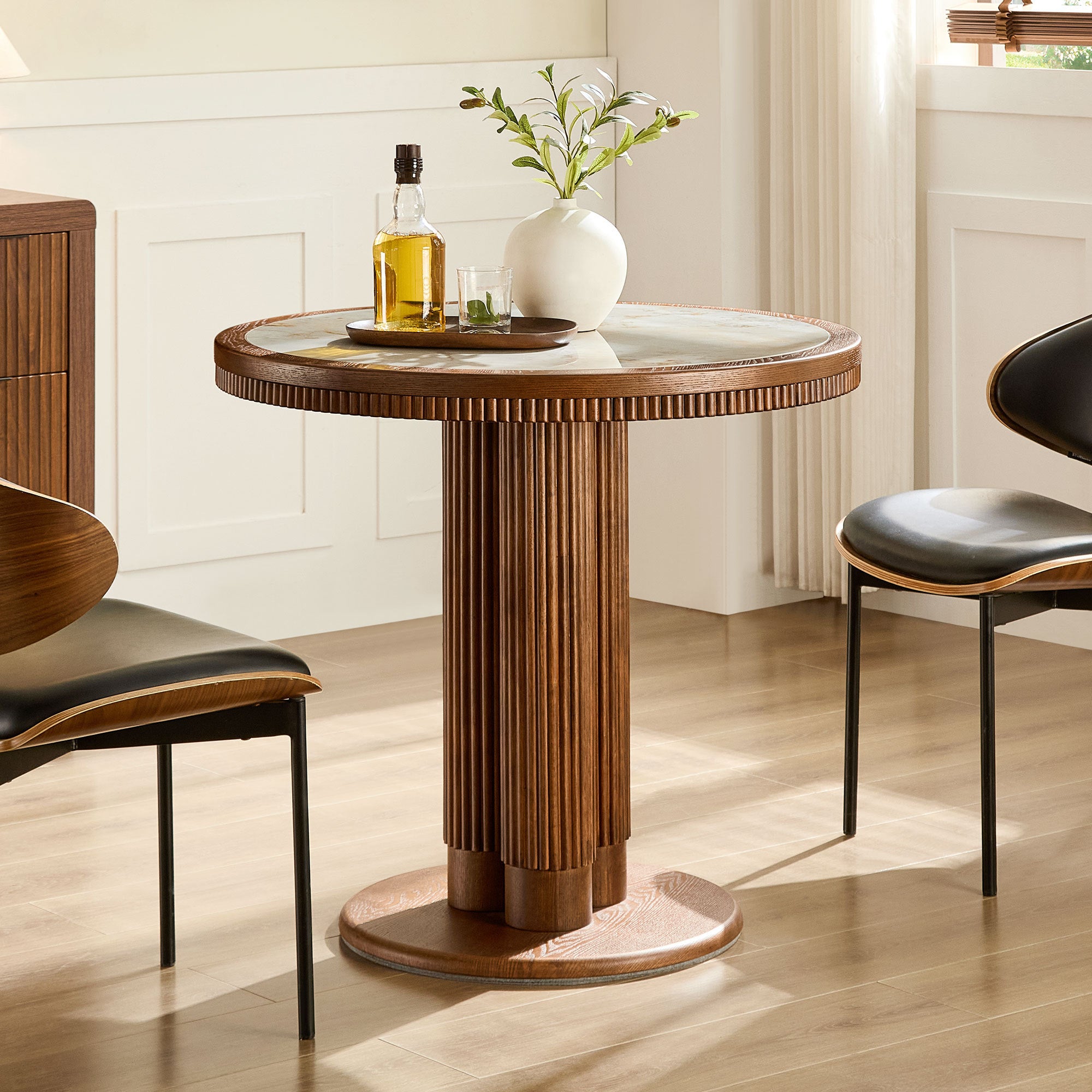 Round walnut dining table with stone top and fluted base beside black chairs in a bright room