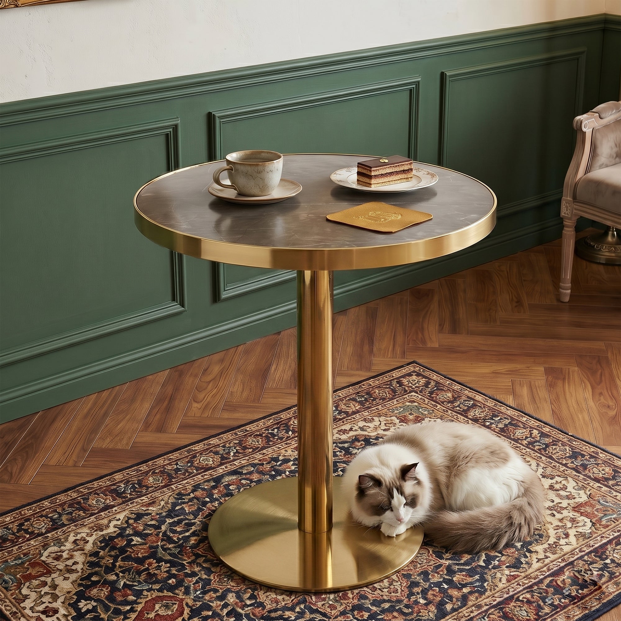Gray faux marble bistro table with gold pedestal base in a green paneled room, cup and cake on top, cat below