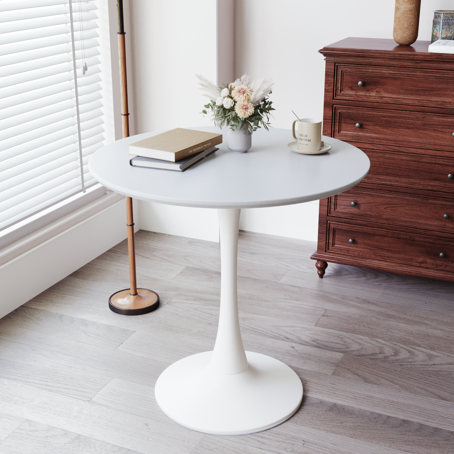 White round tulip dining table styled with books, mug, and flowers by a window beside a wood dresser, three-quarter view