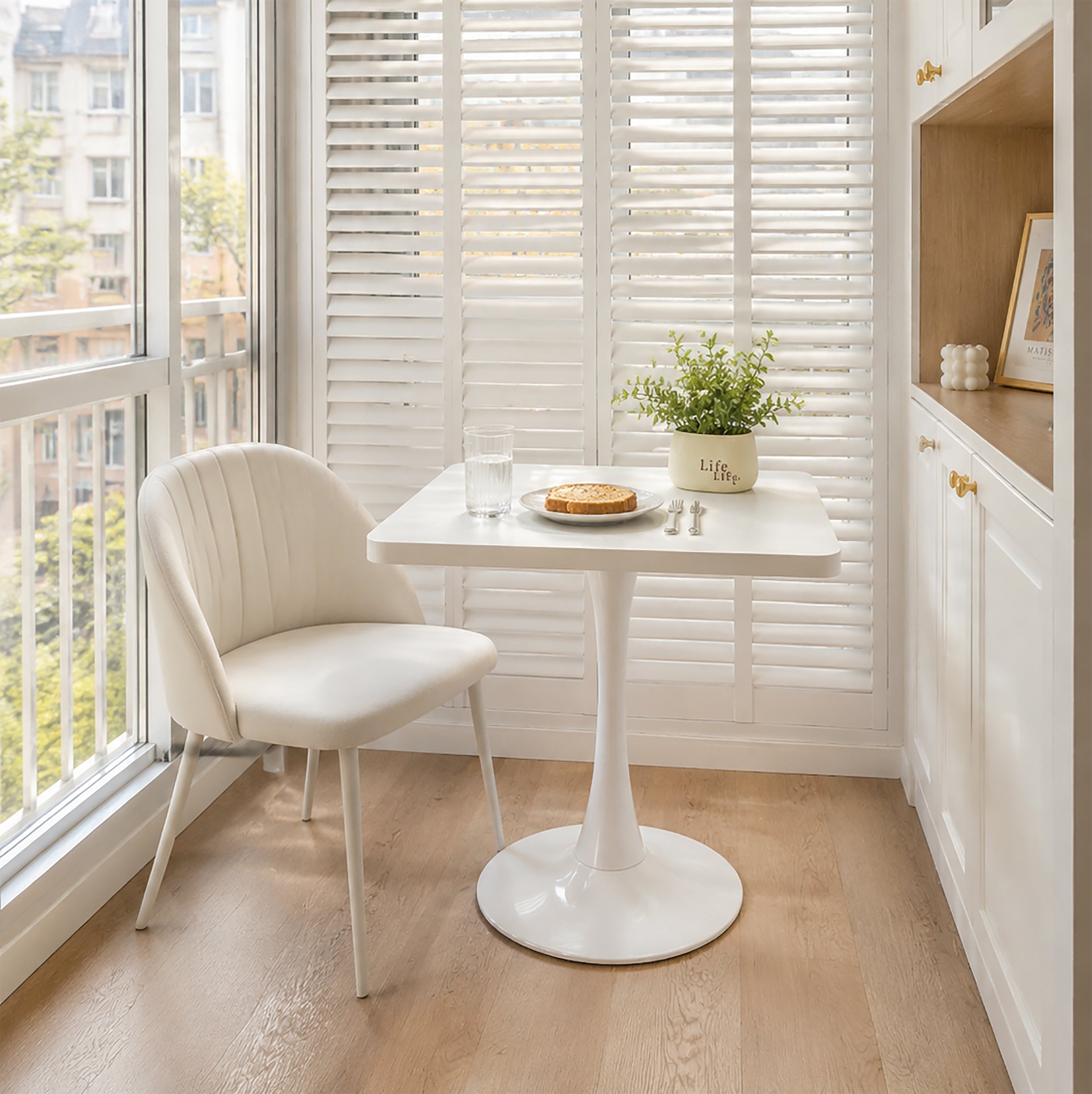 White square tulip bistro table styled in a sunny breakfast nook with a cream chair, plant and plate of toast
