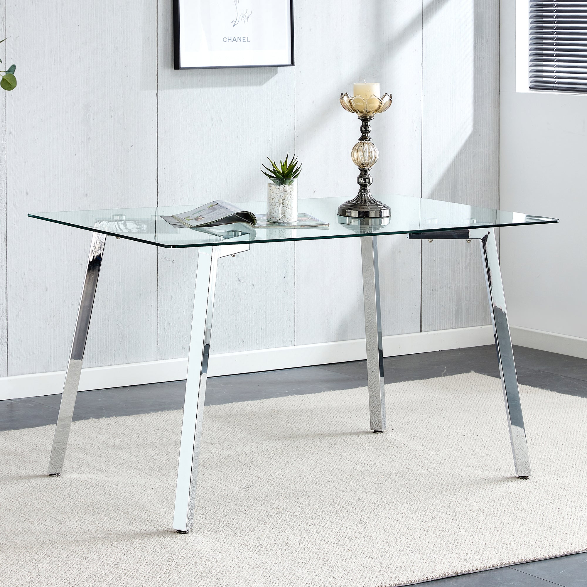 Rectangular glass dining table with chrome legs in a bright room below a framed poster