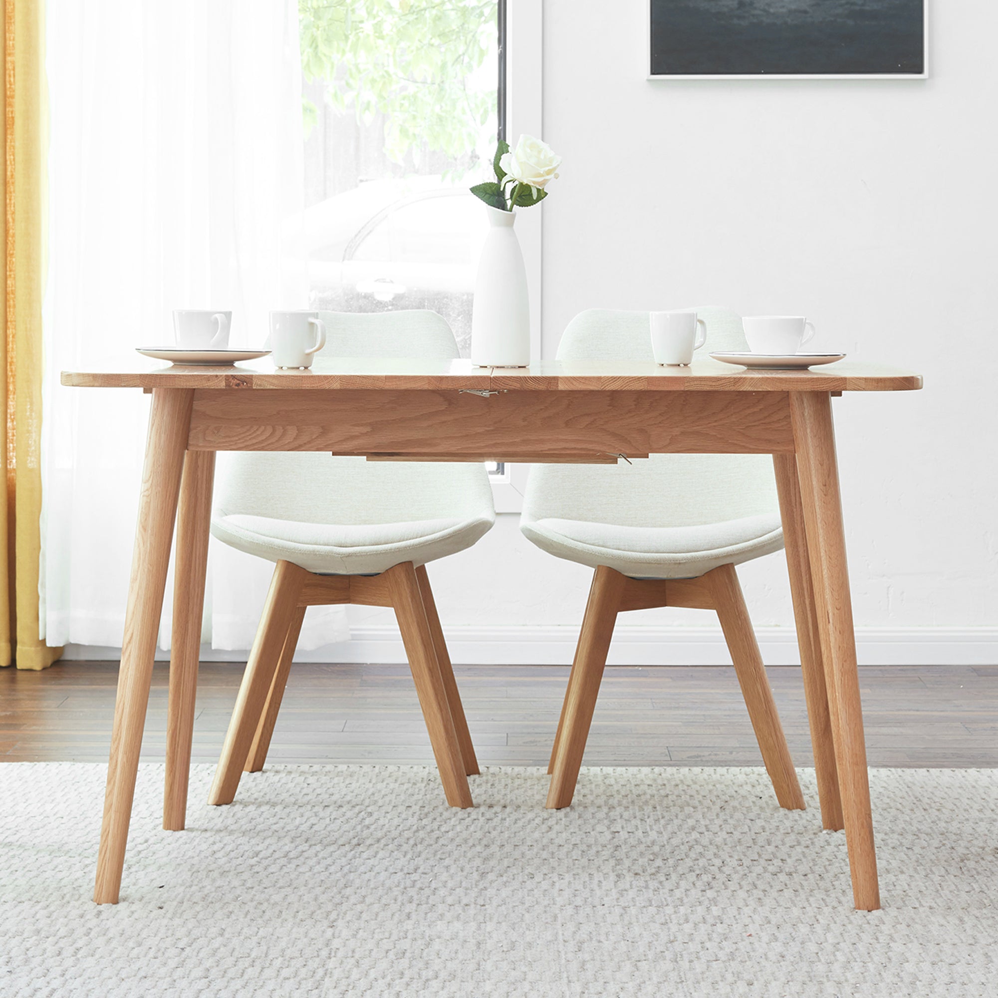 Natural oak dining table with two white chairs and a vase by a sunny window
