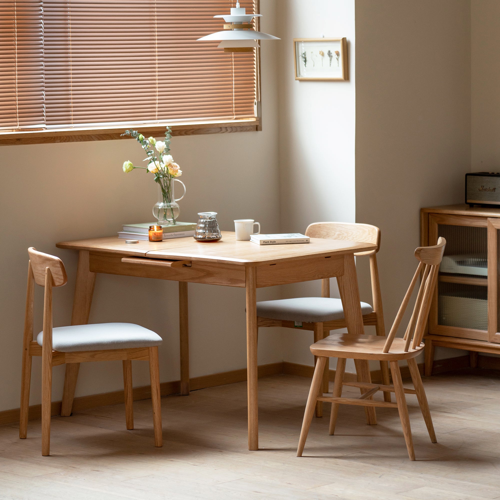 Natural oak dining table with three chairs by a blinded window and pendant lamp, flowers on top
