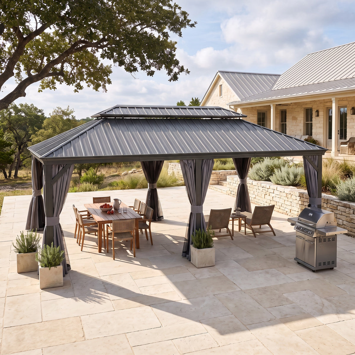 Gray steel double roof hardtop gazebo with tied back curtains over a wood dining table on a stone patio by a house