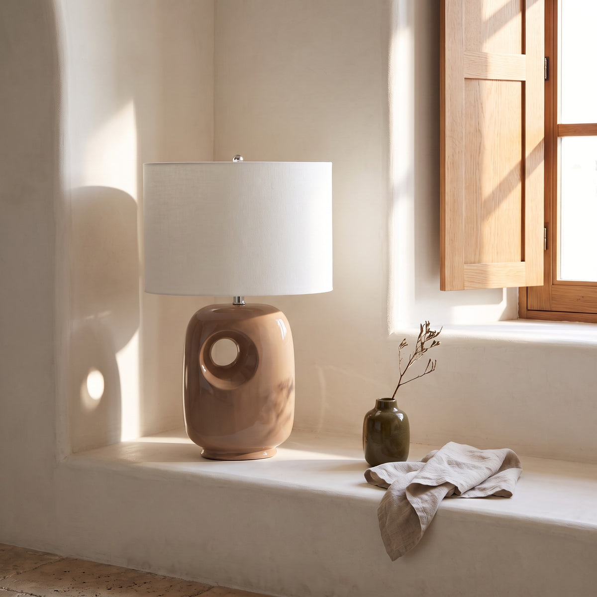 Brown ceramic table lamp with cut-out base and white fabric drum shade on a sunlit plaster window ledge