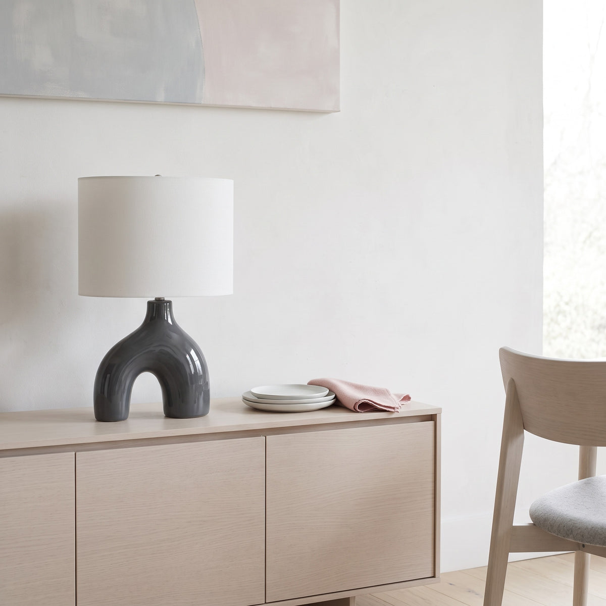 Grey ceramic arch table lamp with ivory drum shade on a light oak sideboard beside white plates