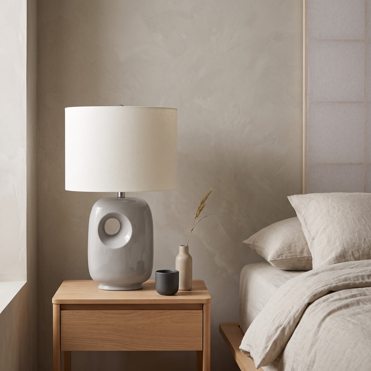 Grey ceramic table lamp with cut-out base and white linen-look drum shade on an oak nightstand beside a bed