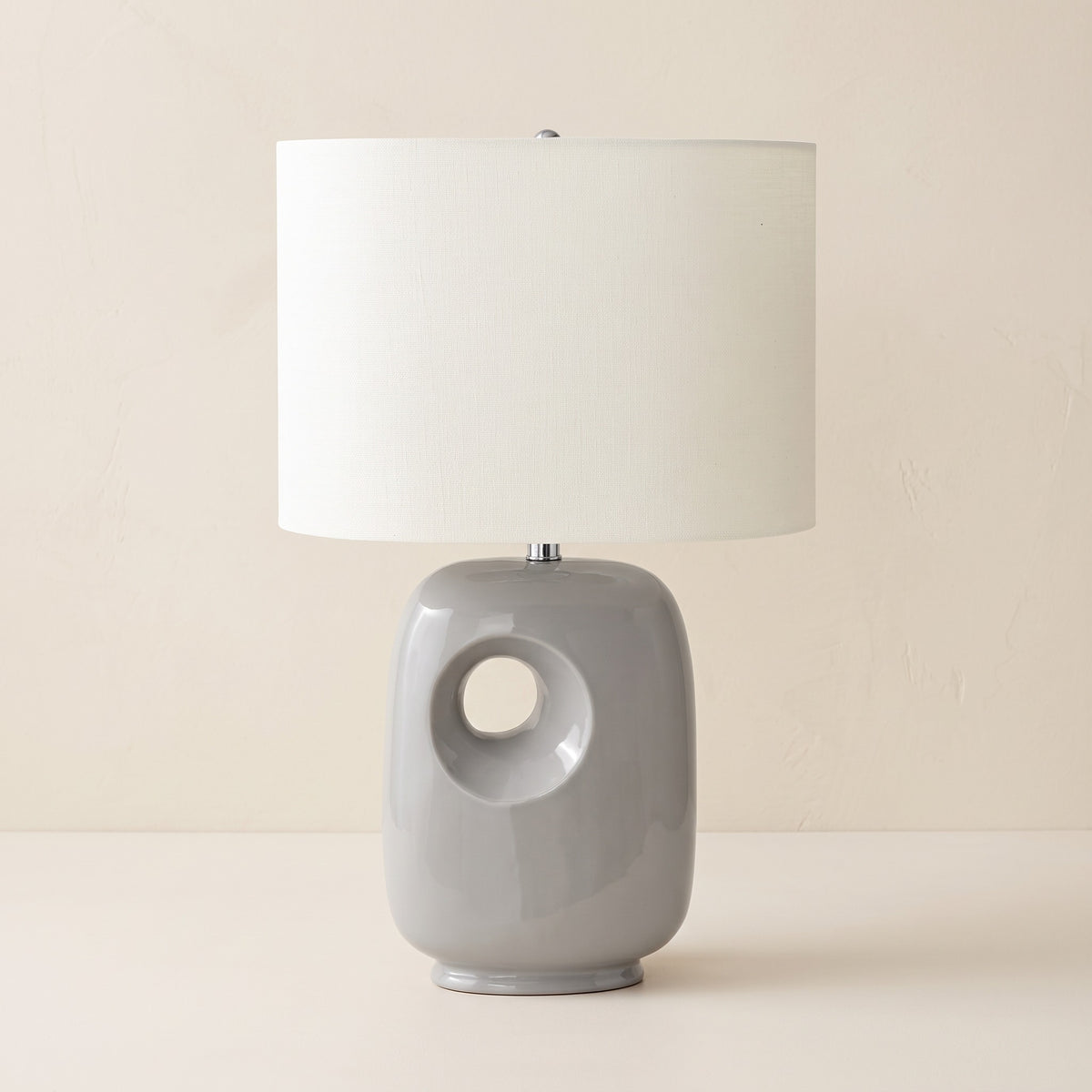 Grey ceramic table lamp with round cut-out base and ivory linen-look drum shade on an ivory plaster backdrop