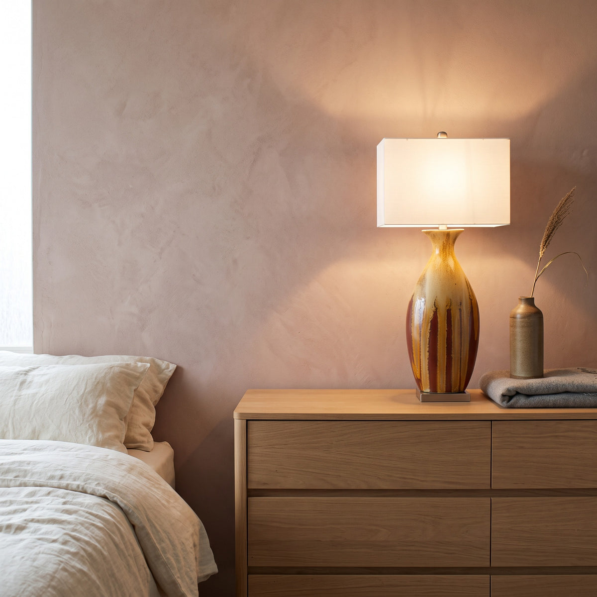 Ceramic table lamp with red and ochre drip glaze and white rectangular shade, lit on an oak dresser beside a bed