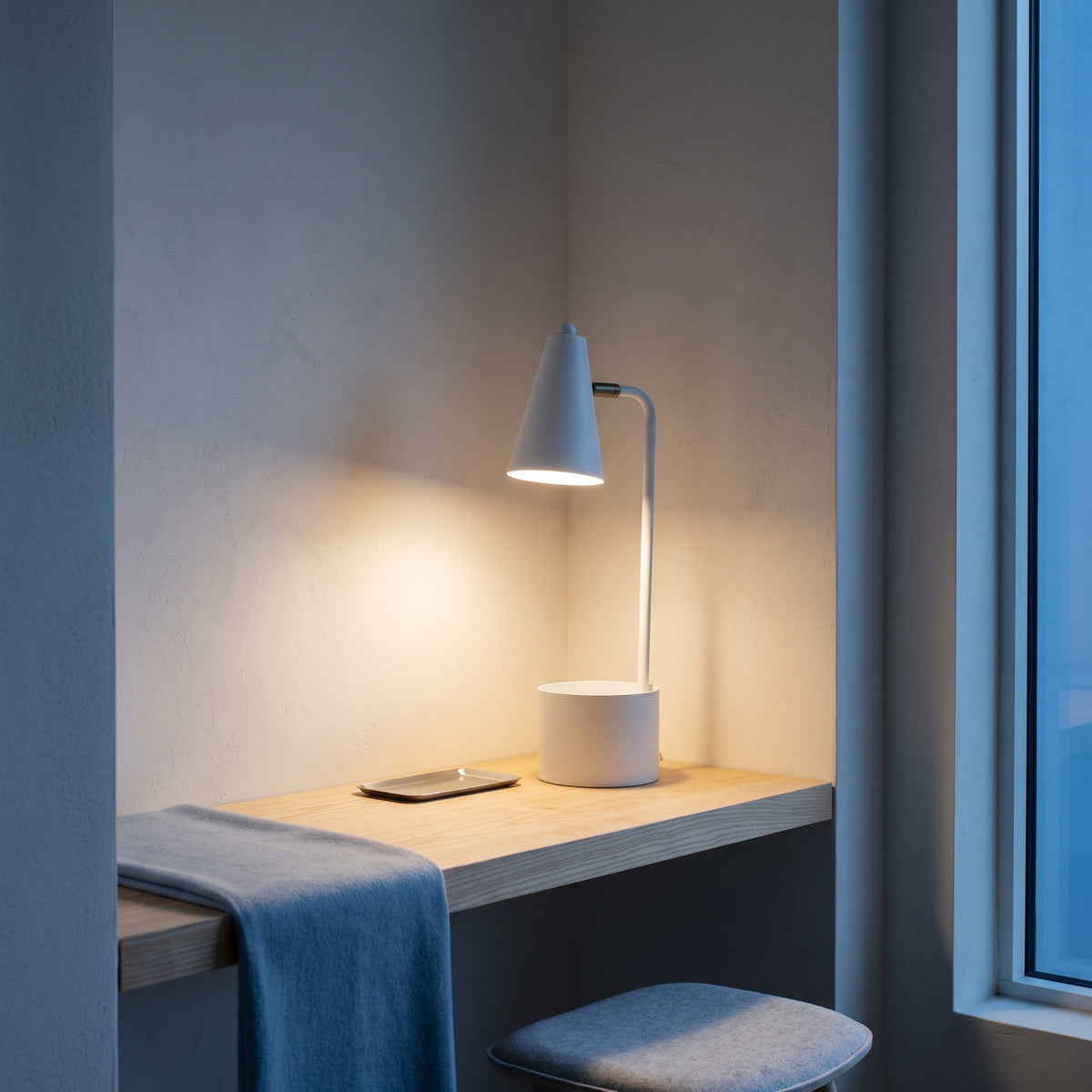 White metal task desk lamp with cone shade and pot base, lit on a wood desk shelf beside a window at dusk