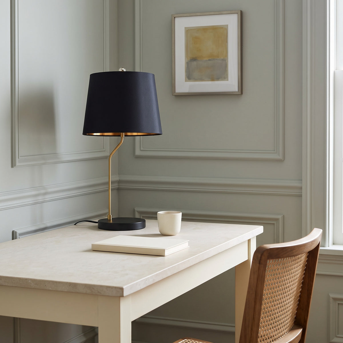 Black table lamp with brass finish stem and black drum shade on a cream desk beside a cane chair