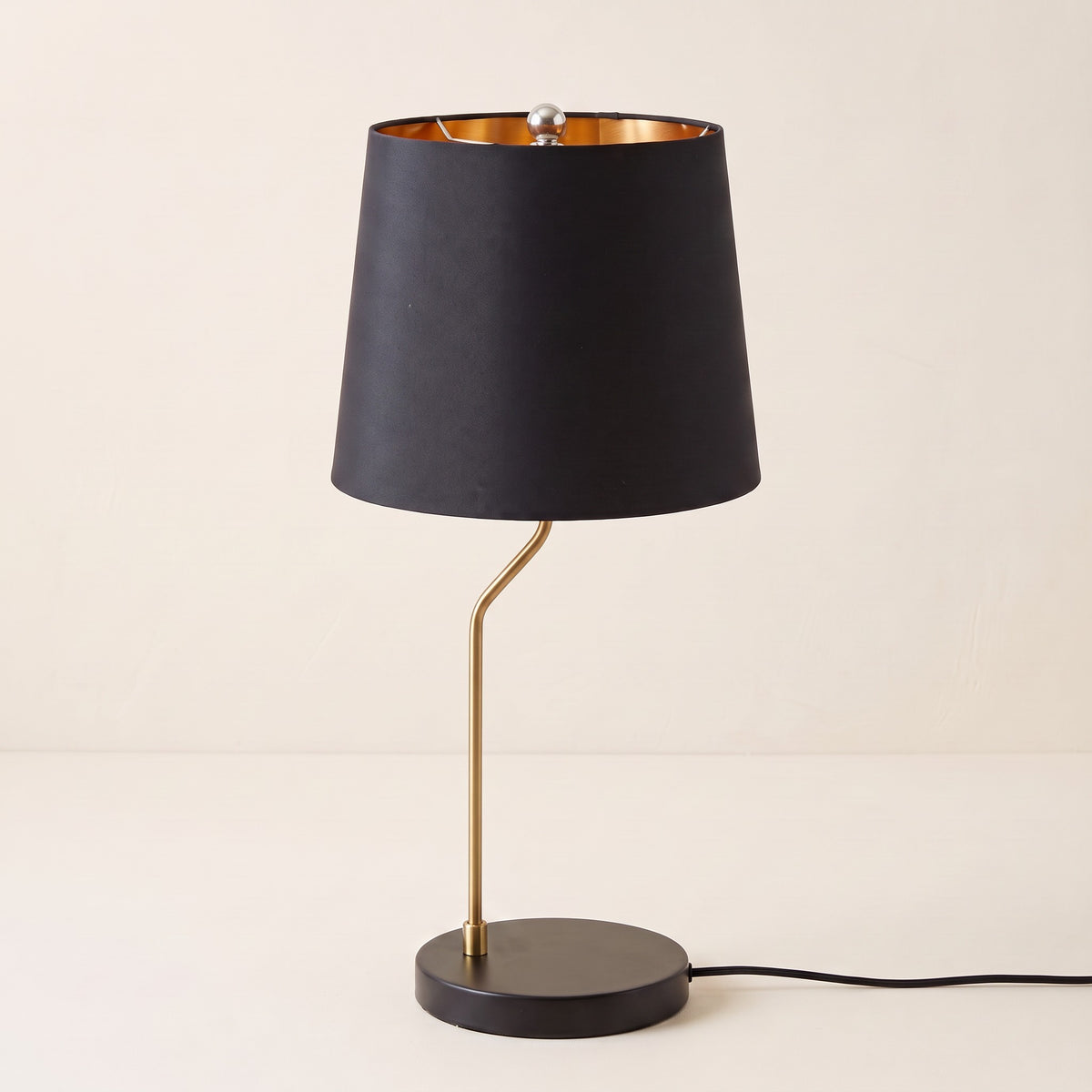 Black table lamp with gold lined drum shade, curved brass stem and round black base on an ivory backdrop