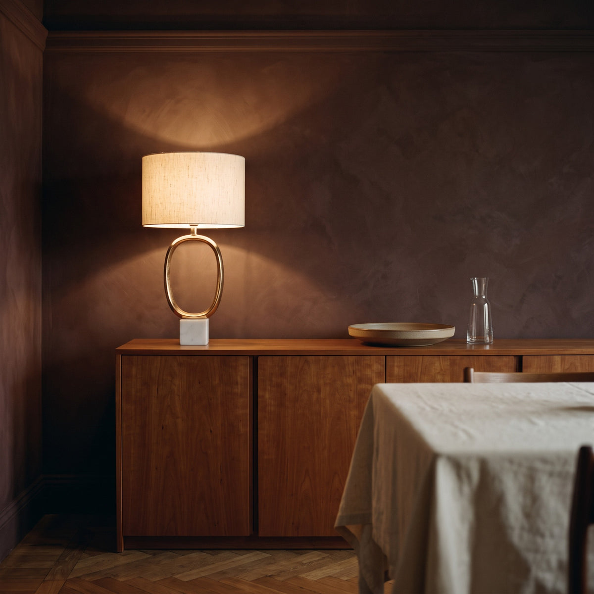 Gold oval cutout table lamp with linen drum shade and white marble base, lit on a wood sideboard in a dining room