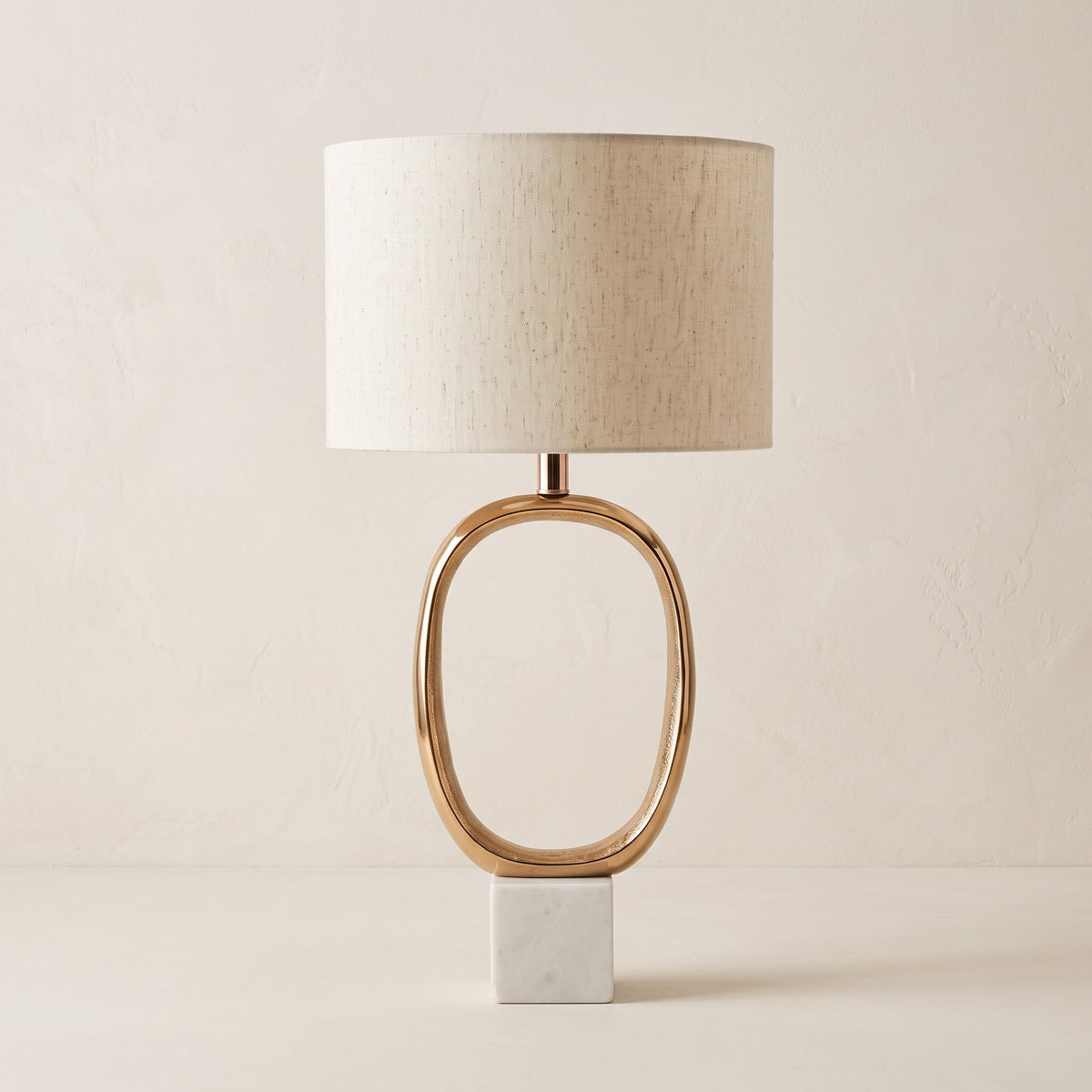 Gold oval cutout table lamp with linen drum shade and white marble block base on an ivory backdrop