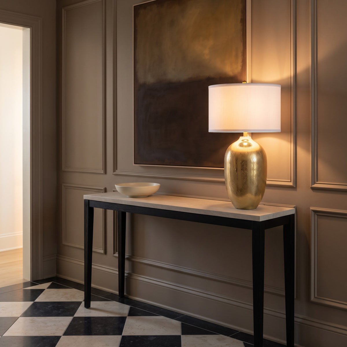 Gold table lamp with crosshatch etched body and white drum shade lit on a stone top console in a panelled entry