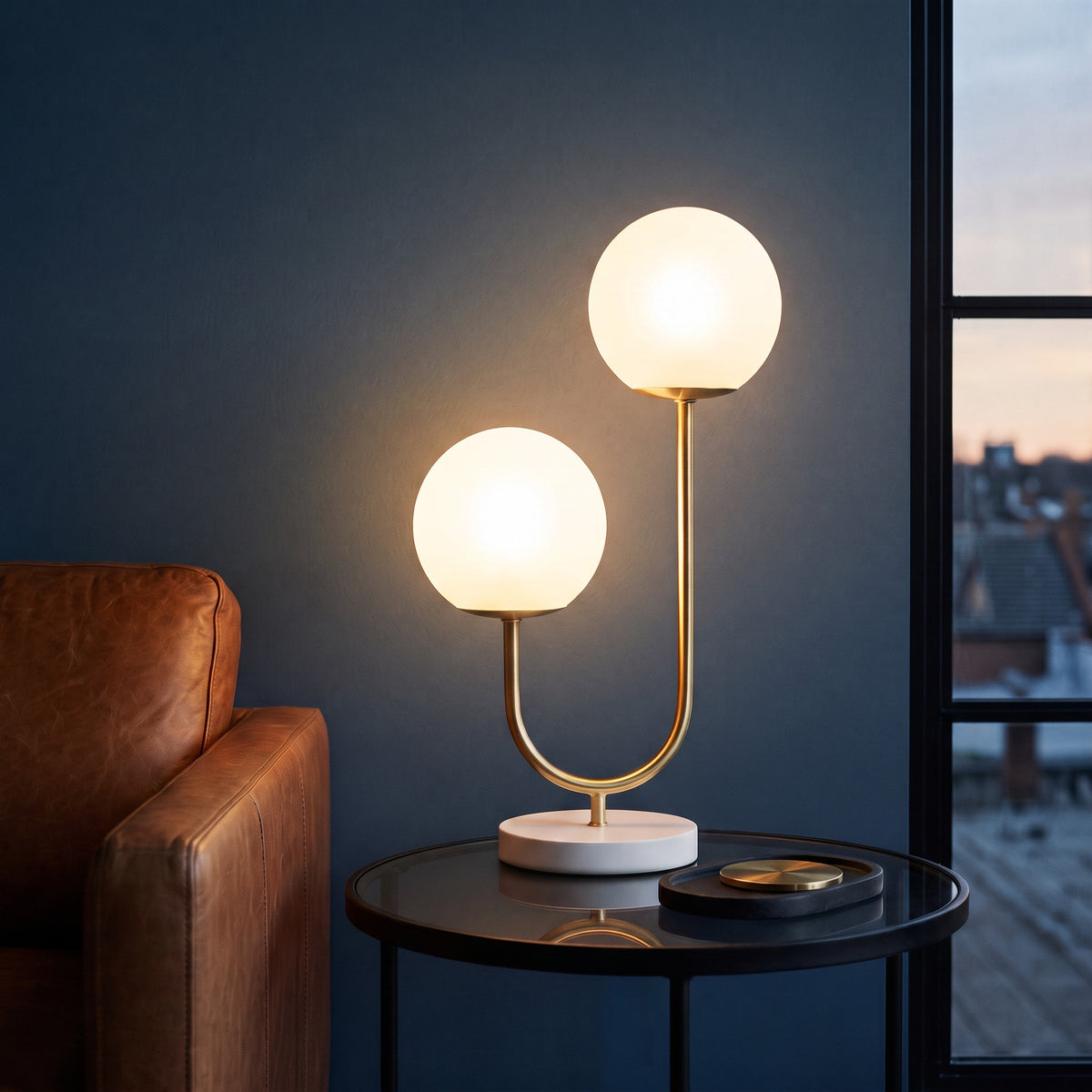 Gold table lamp with two white glass globes lit on a round glass side table beside a leather armchair at dusk