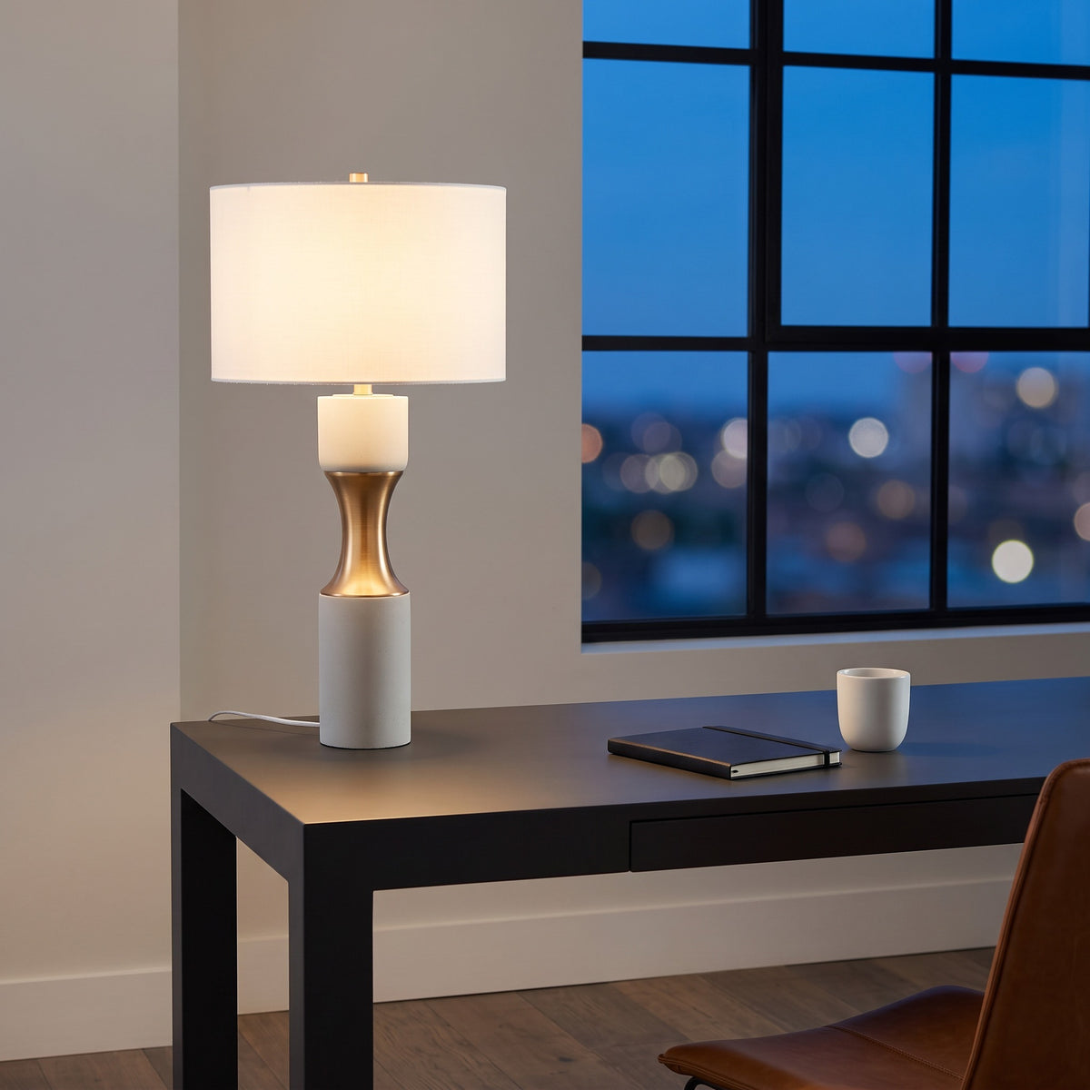White cement table lamp with brass finish waist and white drum shade, lit on a black desk at dusk