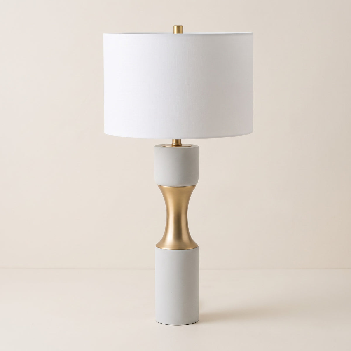 White cement table lamp with brushed brass hourglass waist and white linen drum shade on an ivory backdrop