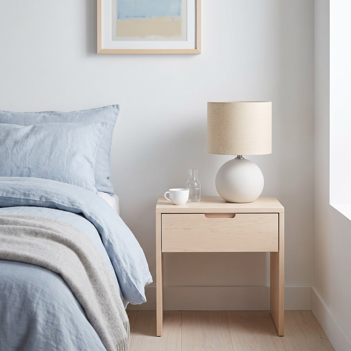 Cream ceramic globe table lamp with beige linen drum shade on a light wood nightstand beside a bed