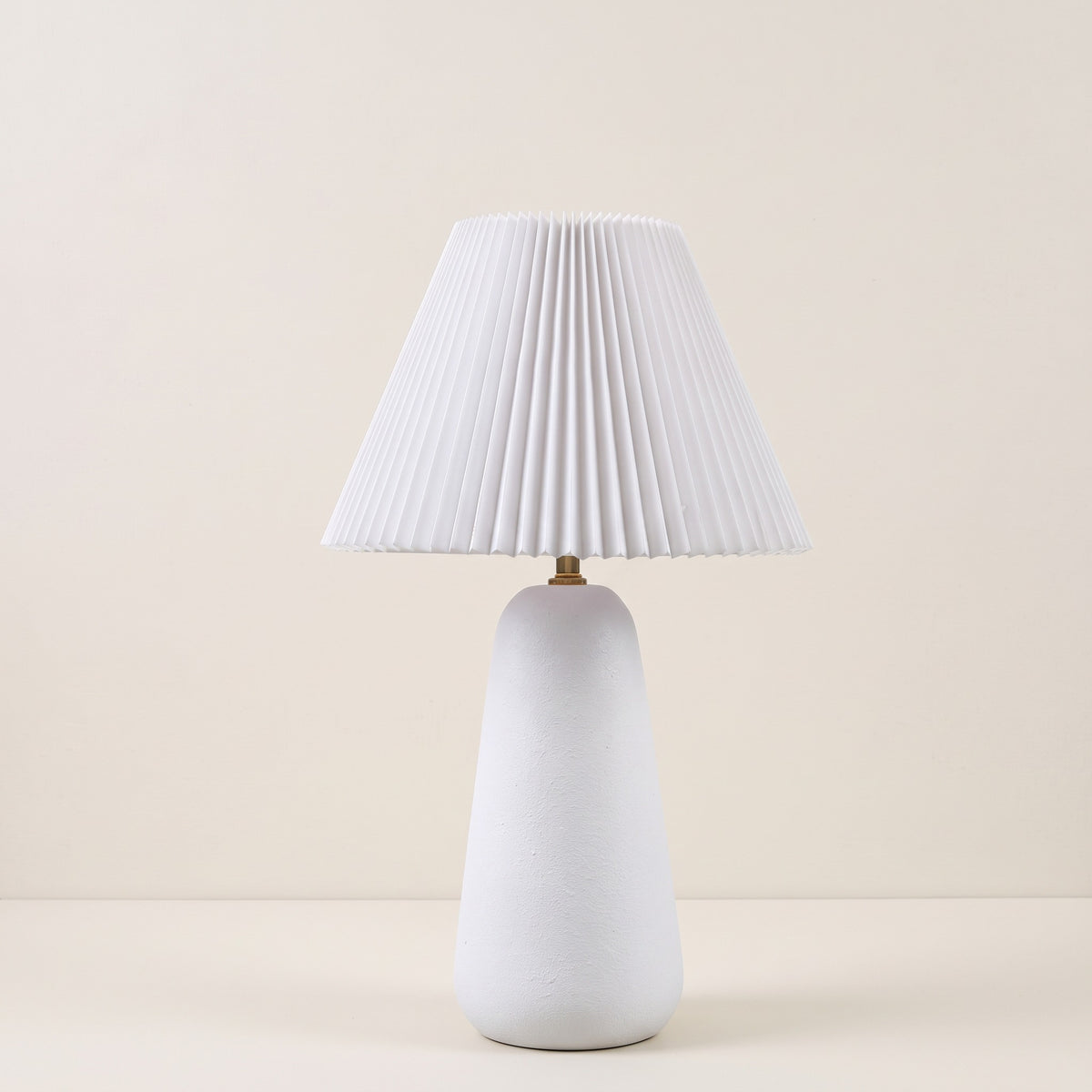 White textured ceramic table lamp with pleated white tapered shade and brass neck on an ivory backdrop