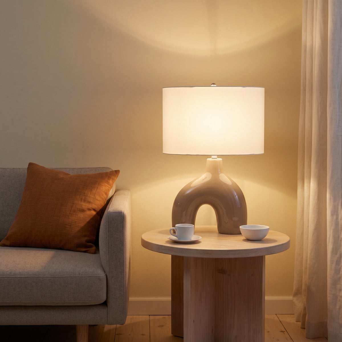 Brown ceramic arch table lamp with ivory drum shade, lit, on a round wood side table beside a sofa