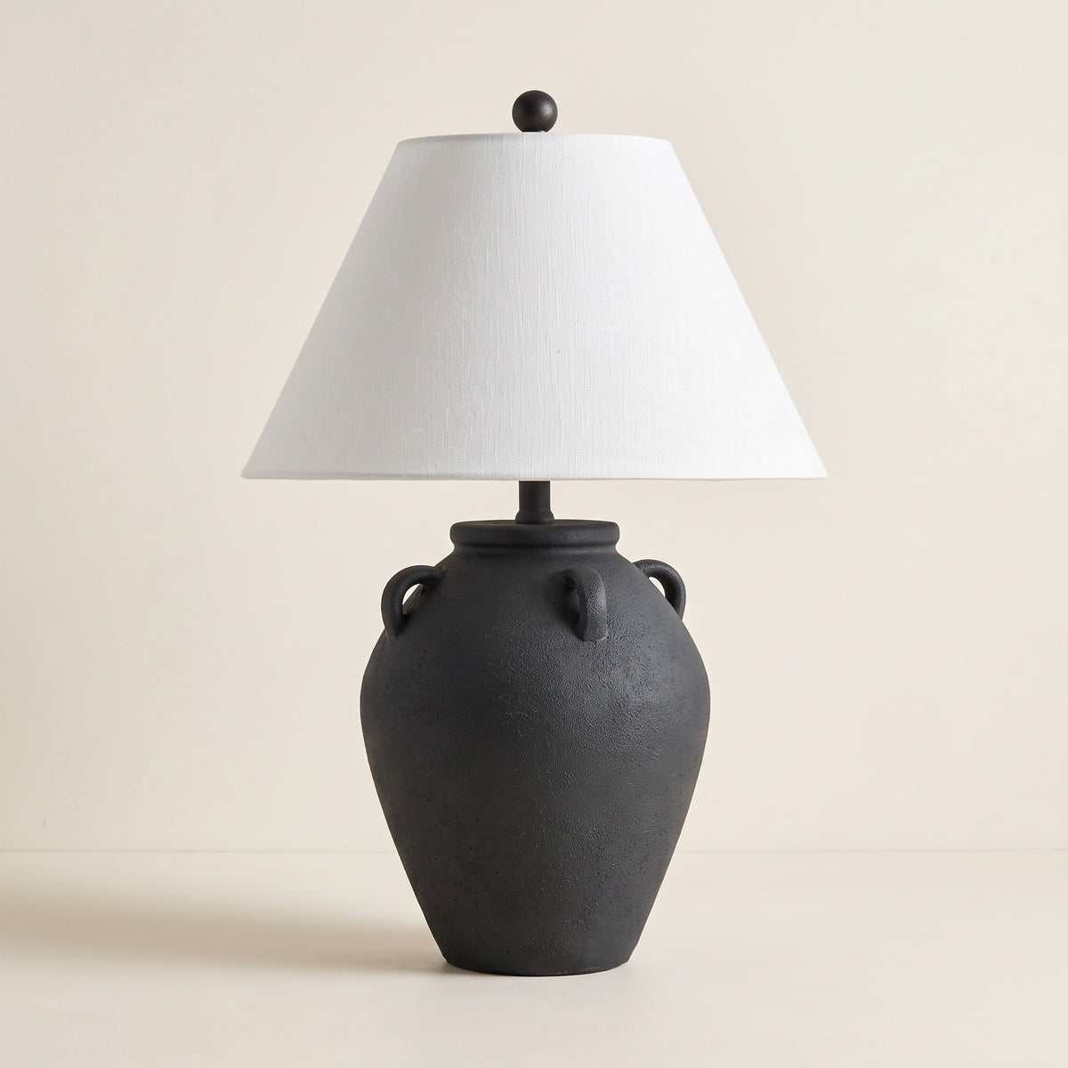 Black textured ceramic table lamp with handles and white linen shade on a warm ivory backdrop
