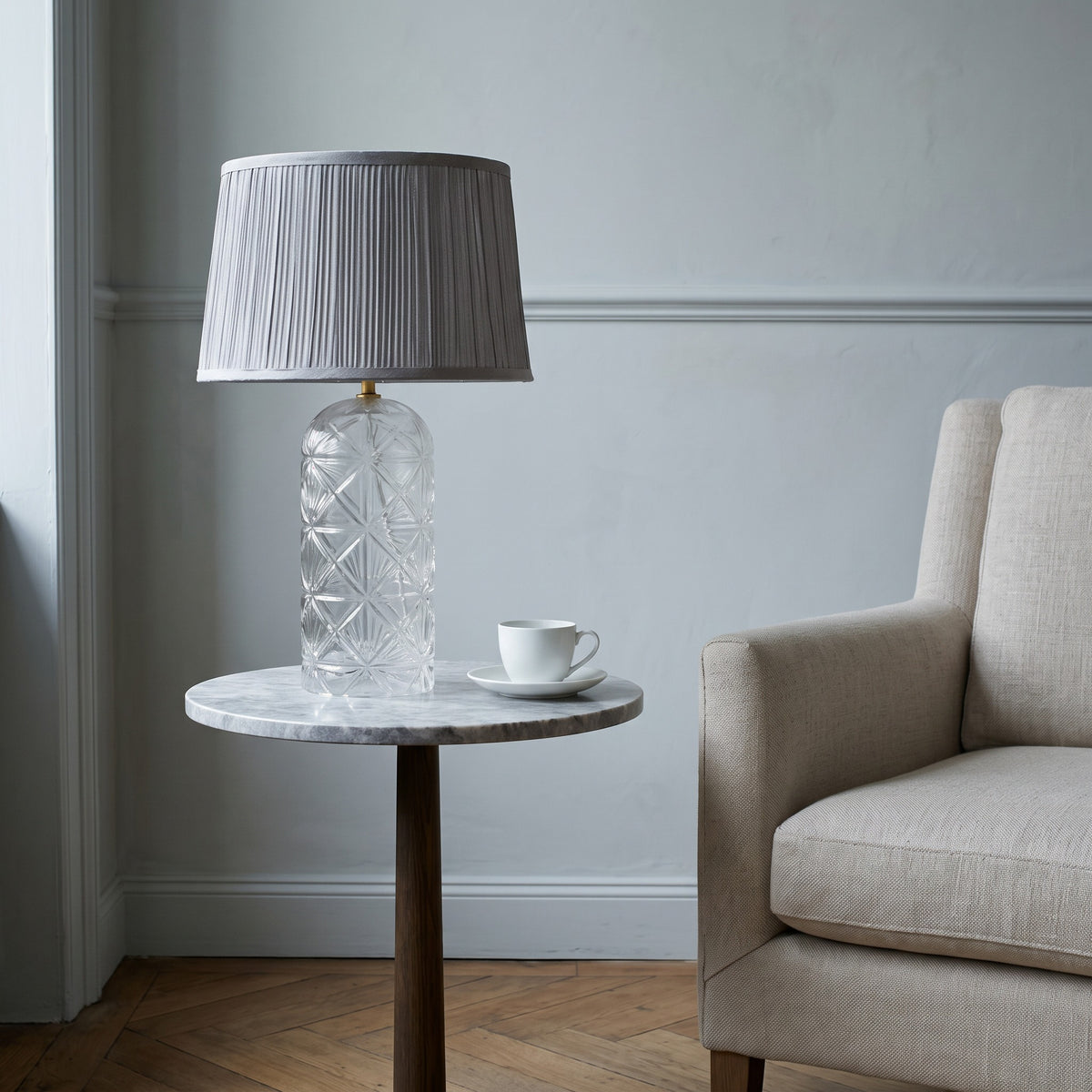 Clear cut glass table lamp with pleated grey fabric shade on a marble side table beside a linen armchair
