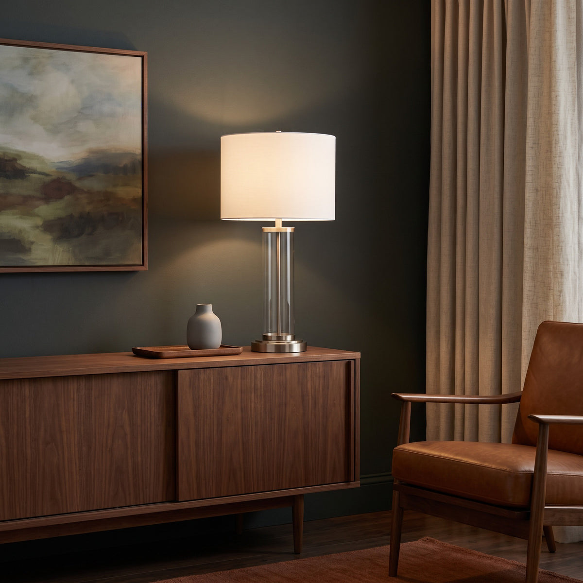 Clear glass table lamp with brushed nickel base and lit white drum shade on a walnut sideboard