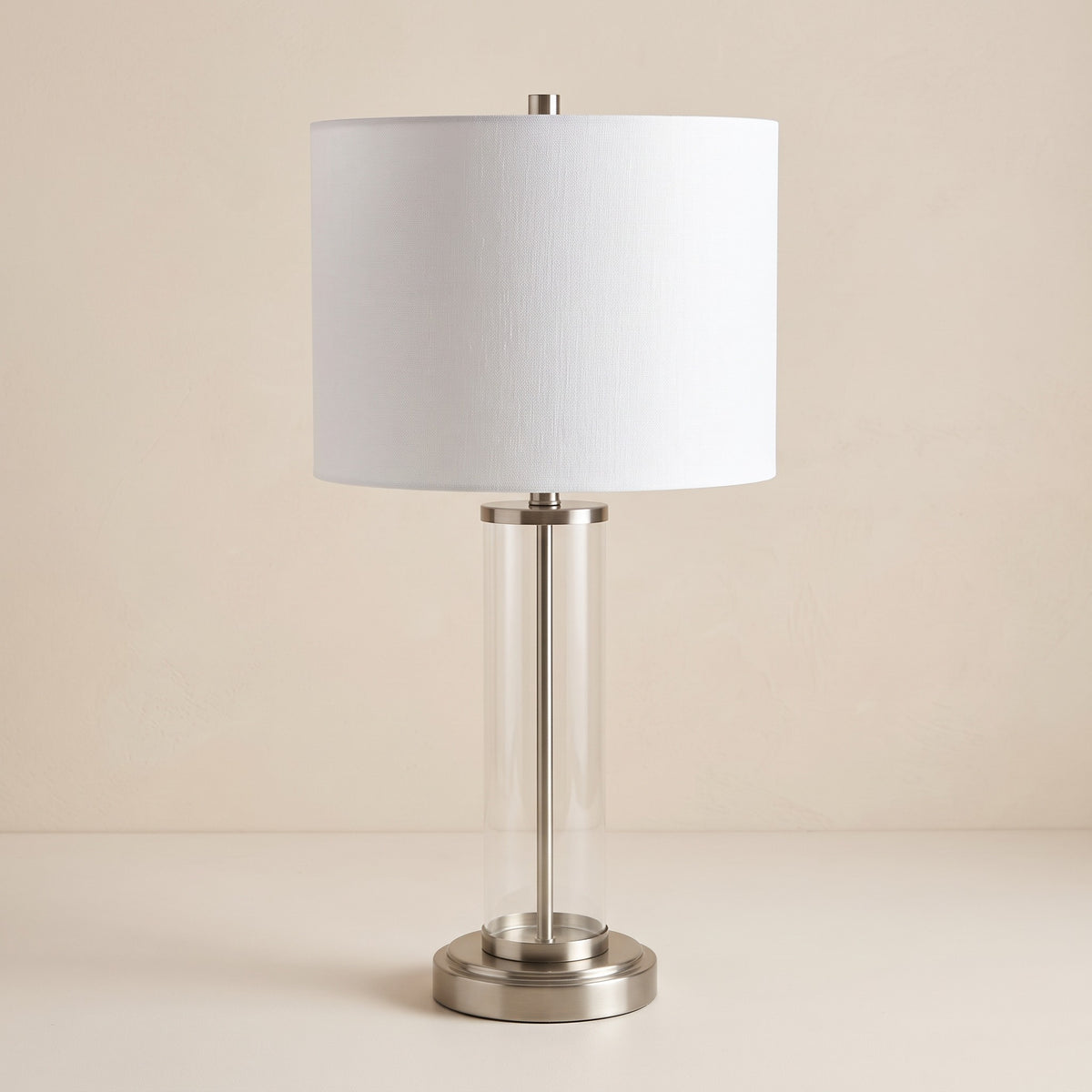 Brushed nickel table lamp with clear glass column and white linen-look drum shade on an ivory backdrop