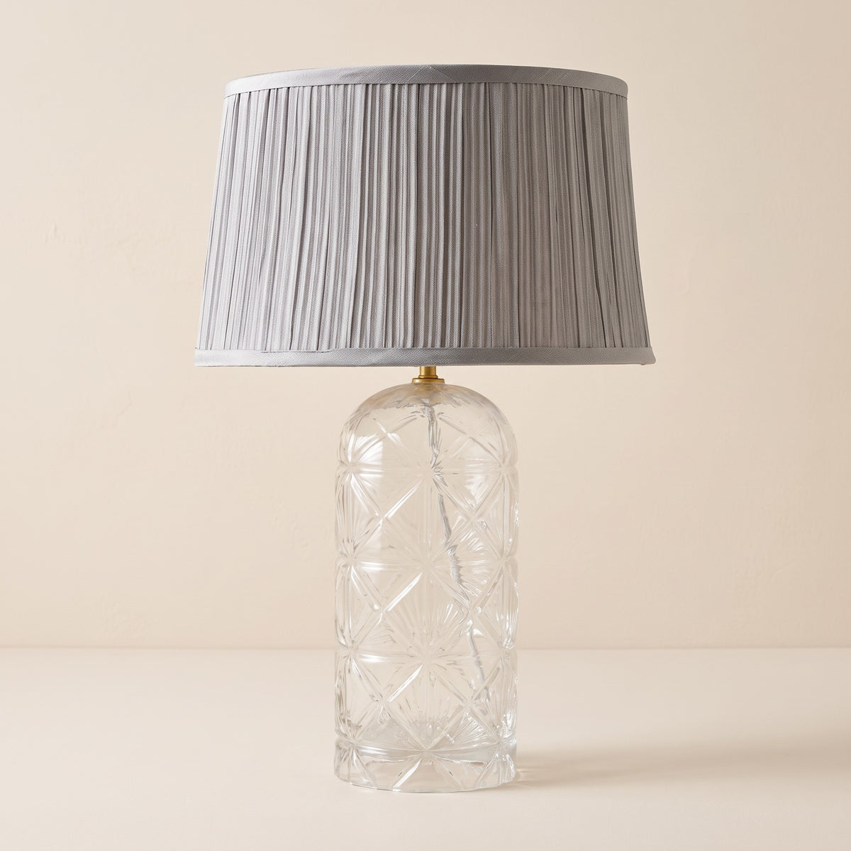Clear cut glass table lamp with pleated grey fabric shade and gold fitting on an ivory backdrop