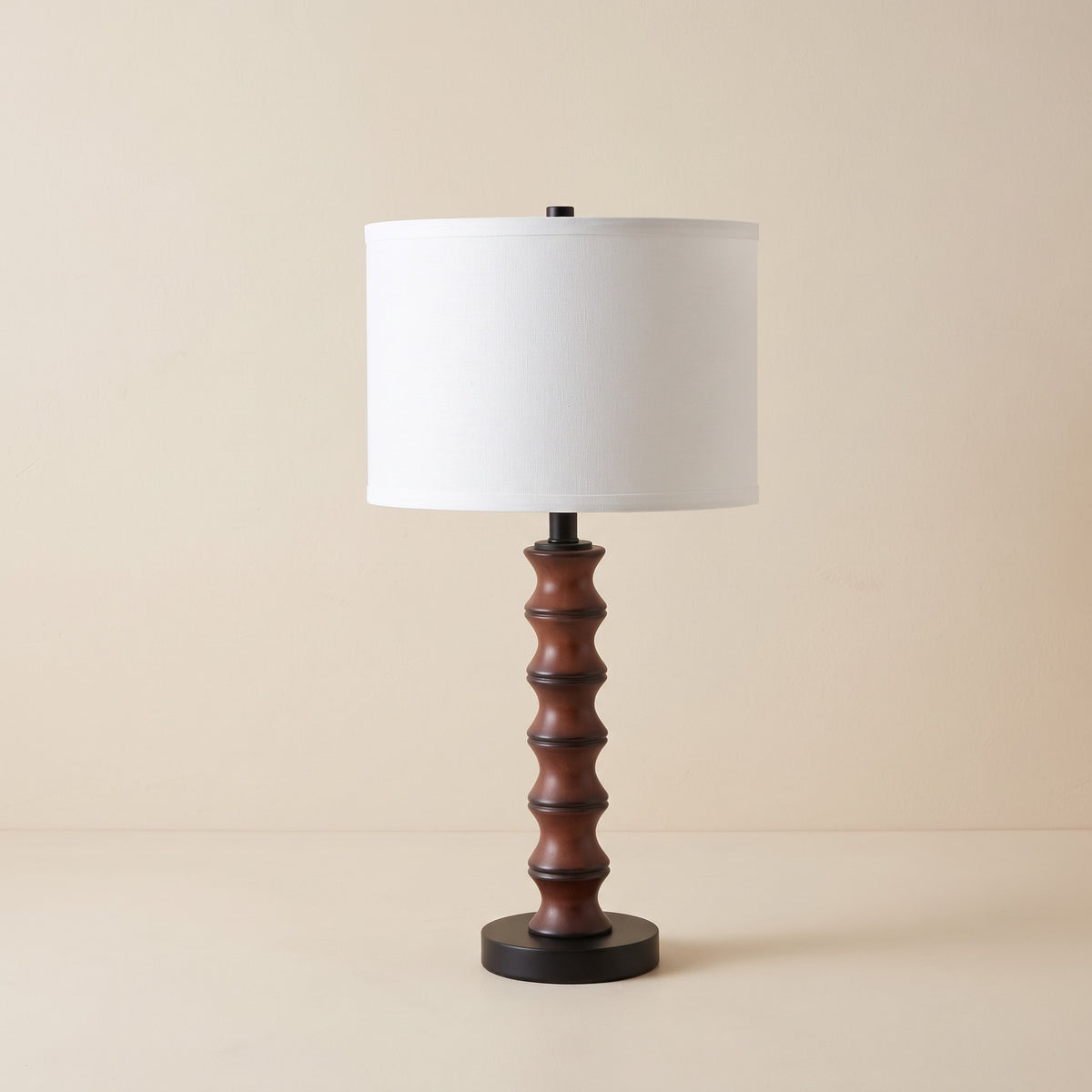 Cherry brown turned table lamp with white linen-look drum shade and black round base on an ivory backdrop