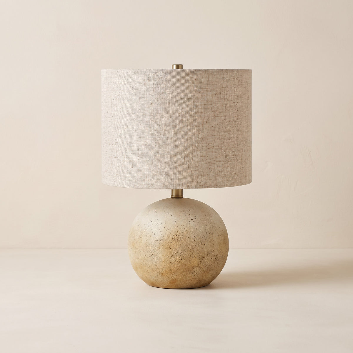 Beige concrete sphere table lamp with linen drum shade and brass finial on an ivory plaster backdrop