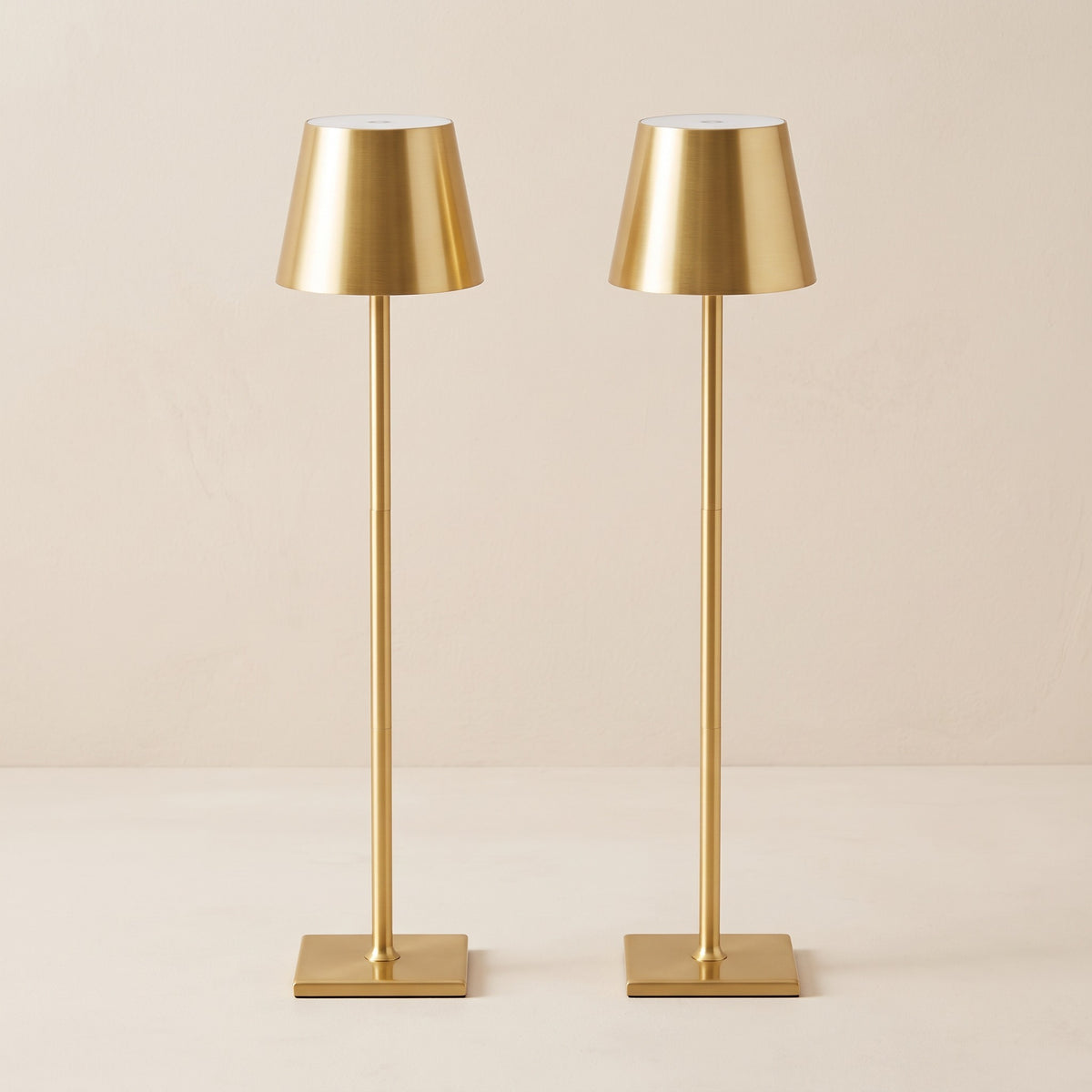 Two gold cordless table lamps with tapered metal shades and square bases on an ivory backdrop