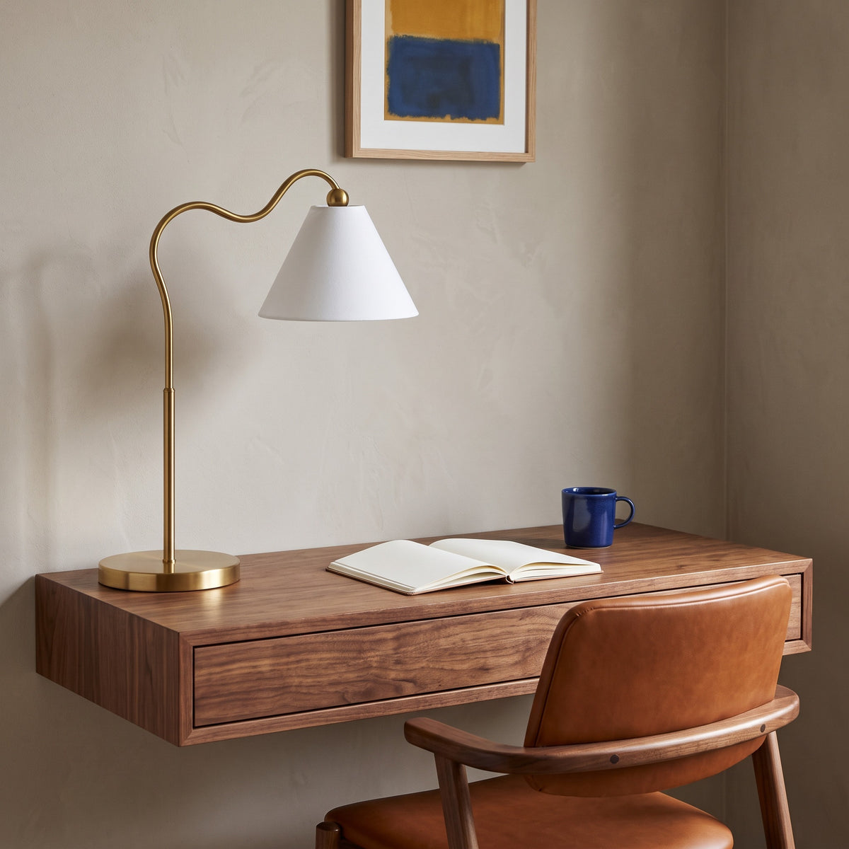 Gold table lamp with curved arm and white fabric cone shade on a floating walnut desk