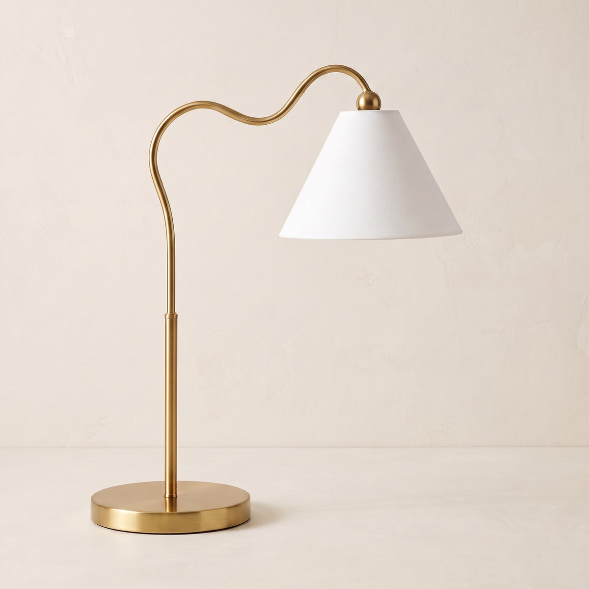 Gold table lamp with curved wavy arm and white cone fabric shade on an ivory plaster backdrop