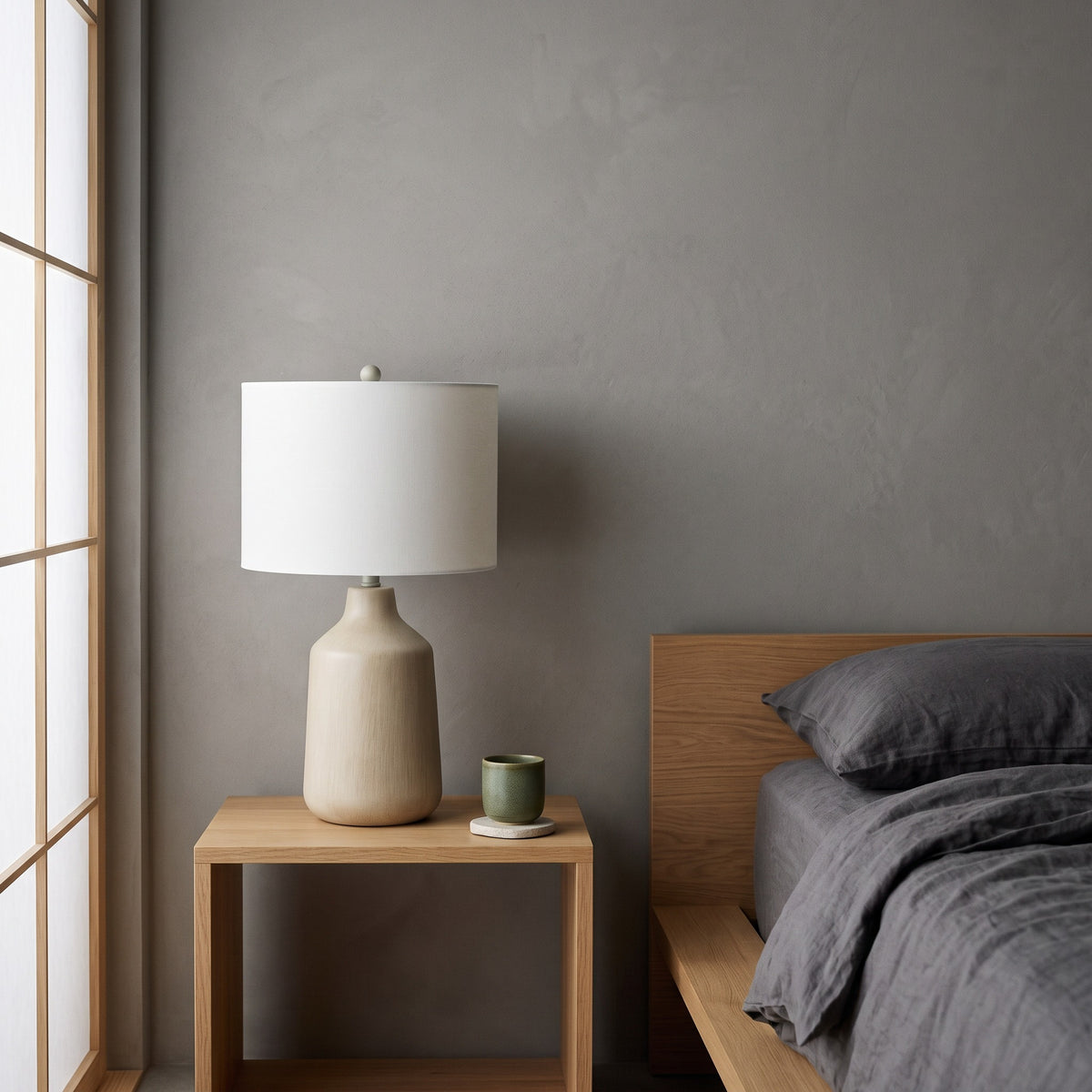 Beige concrete table lamp with white drum shade on an oak nightstand beside a bed with gray linen