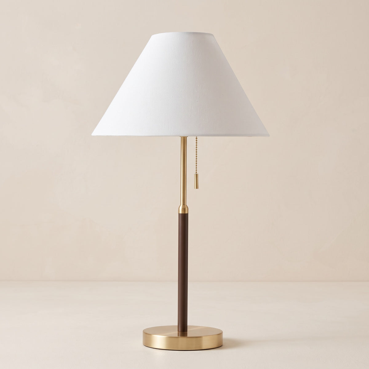 Table lamp with white tapered fabric shade, brass and dark brown stem and pull chain on an ivory backdrop