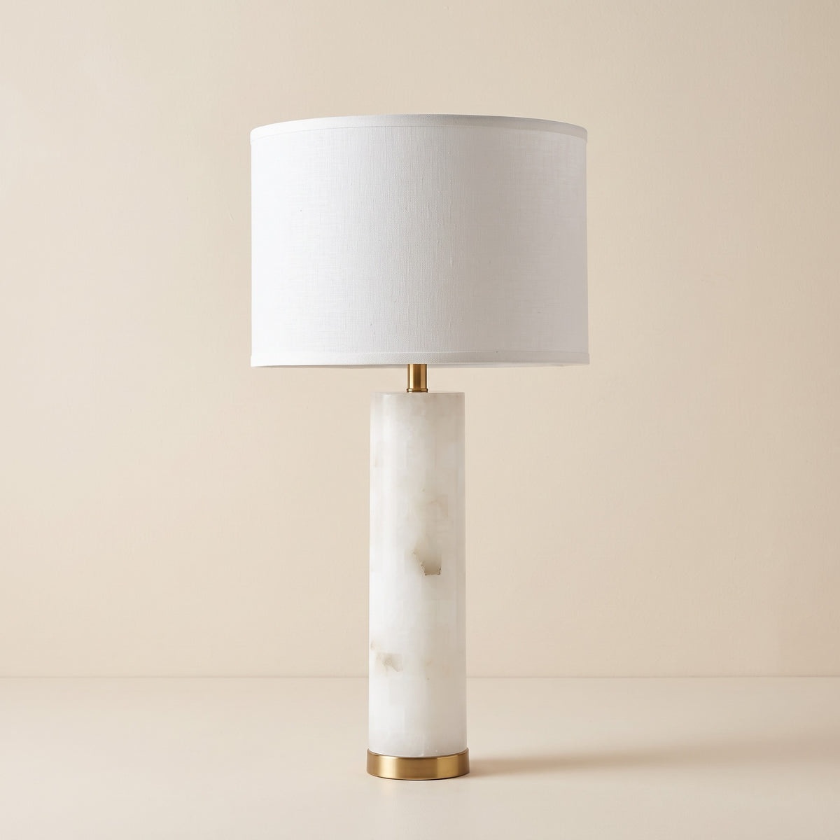 White alabaster column table lamp with gold base ring and white linen drum shade on an ivory backdrop