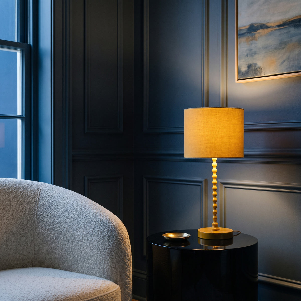 Yellow table lamp with linen-look drum shade, lit, on a black round side table beside a cream boucle chair