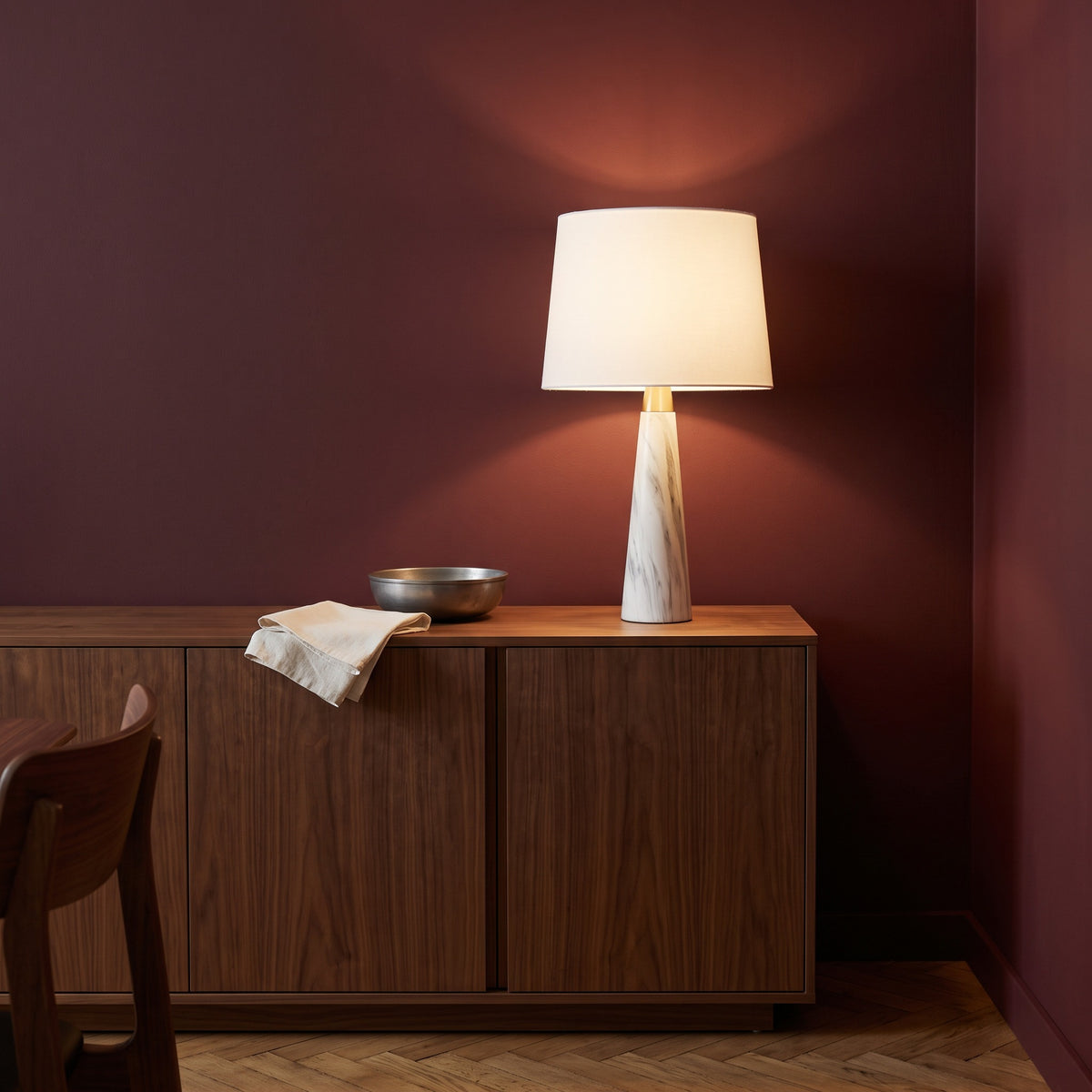 White marble-look table lamp with white fabric shade, lit, on a walnut sideboard against a burgundy wall