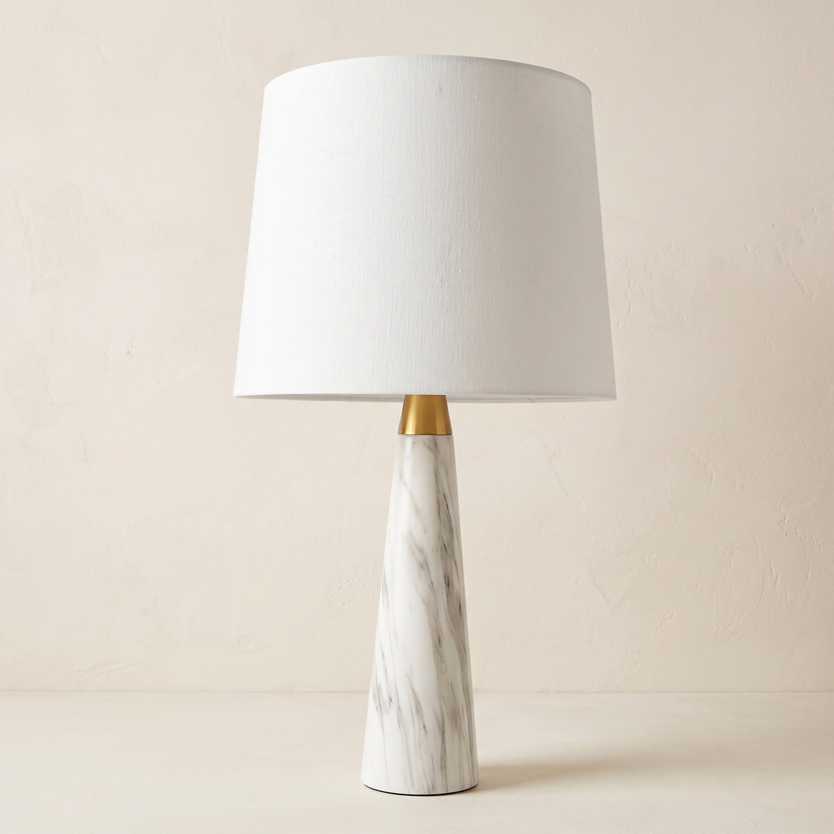 White marble-look table lamp with brass finish collar and white fabric drum shade on an ivory plaster backdrop