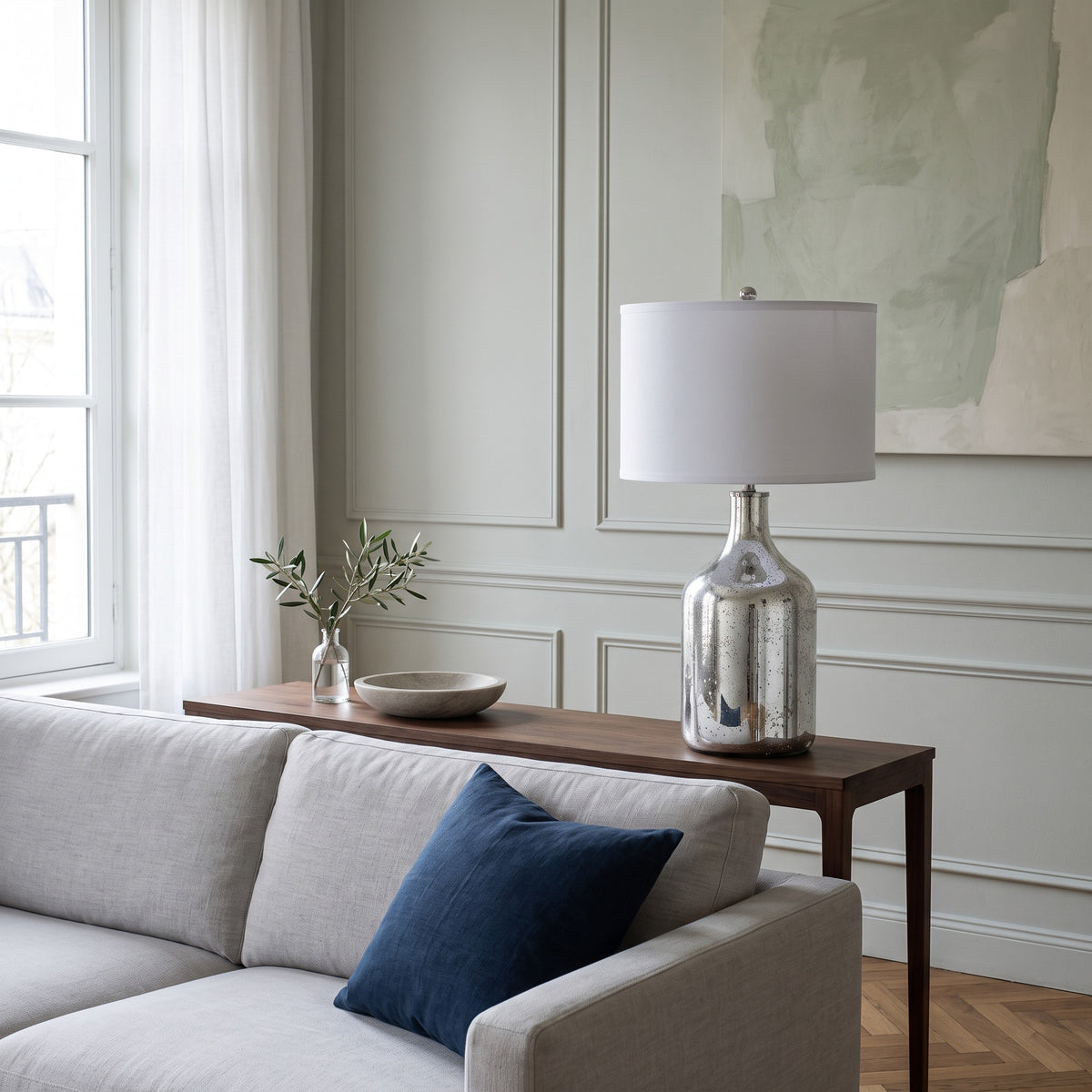 Silver mercury glass table lamp with white drum shade on a walnut console behind a light gray sofa