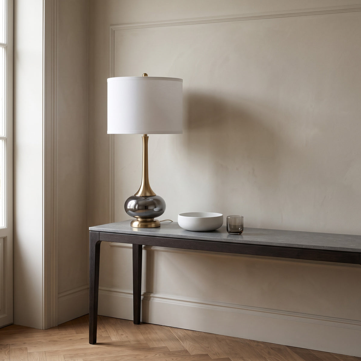 Gray glass table lamp with gold neck and white drum shade on a gray stone top console by a window